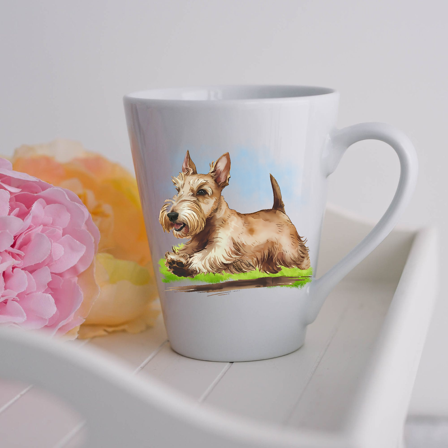 A Wheaten Scottish Terrier is seen on a white ceramic latte mug running on a grassy surface.  The mug is placed on a white tray with pink and yellow flowers. 