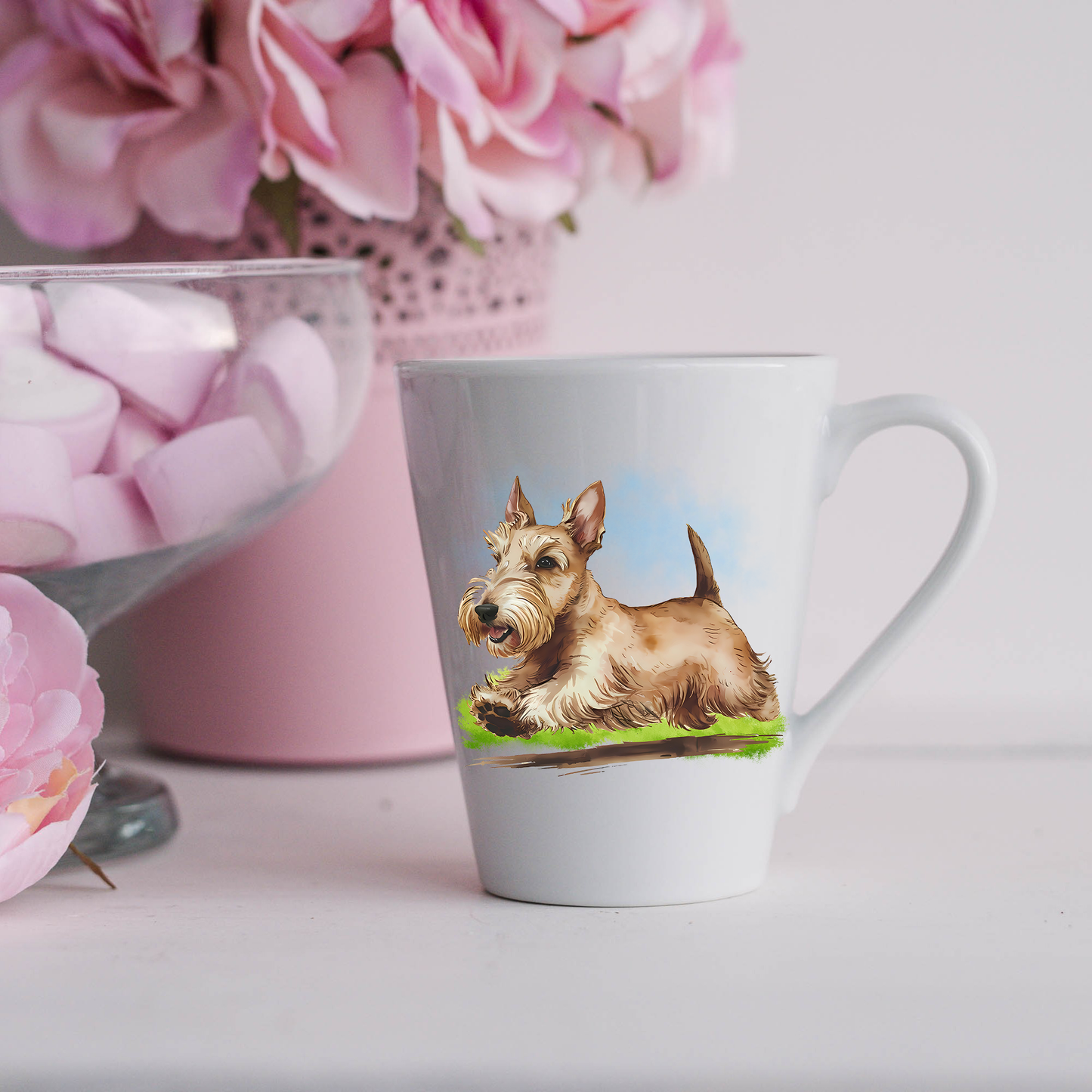 A Wheaten Scottish Terrier is running on a grassy surface on a white ceramic latte mug. The mug is placed on a white surface with pink flowers in a pink plant pot and a dish of pink marshmallows.