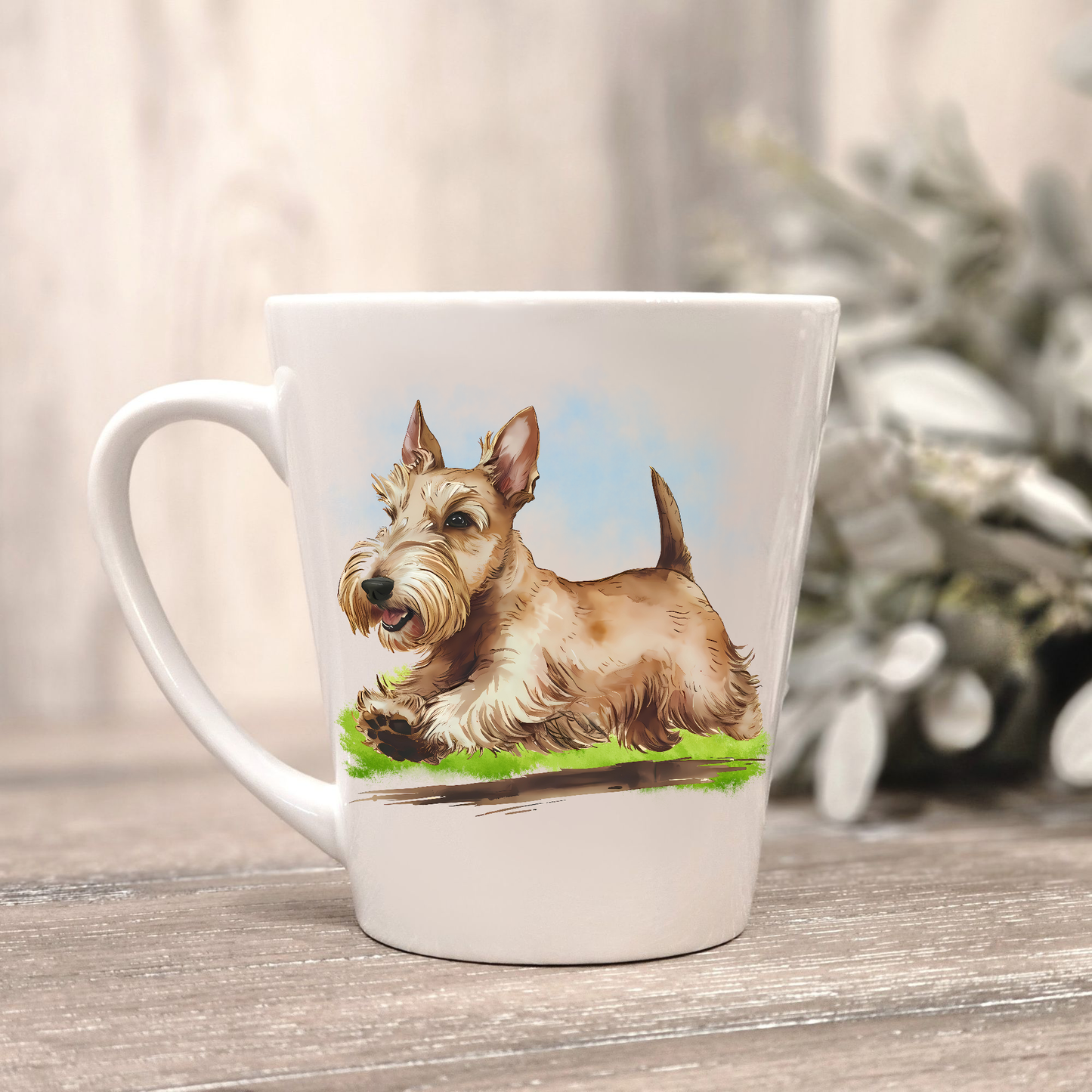 A Wheaten Scottish Terrier is running on a grassy surface on a white ceramic latte mug. The mug is placed on a wooden surface with sage green foliage to the right-hand side.