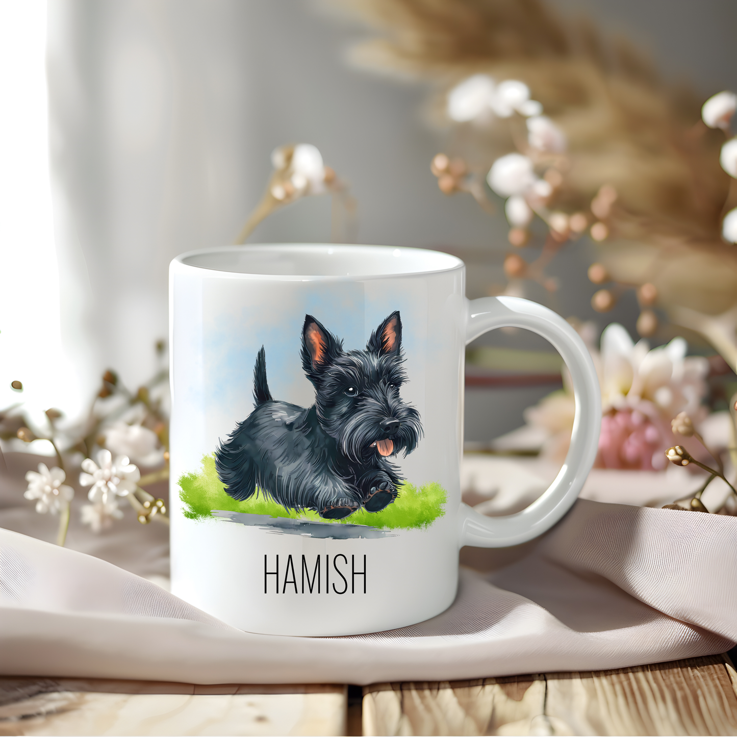 A lovely black Scottish Terrier coffee mug personalised with a pet name.  The mug is sat on a wooden surface with a creamy coloured cloth and a variety of flowers decorate the background.