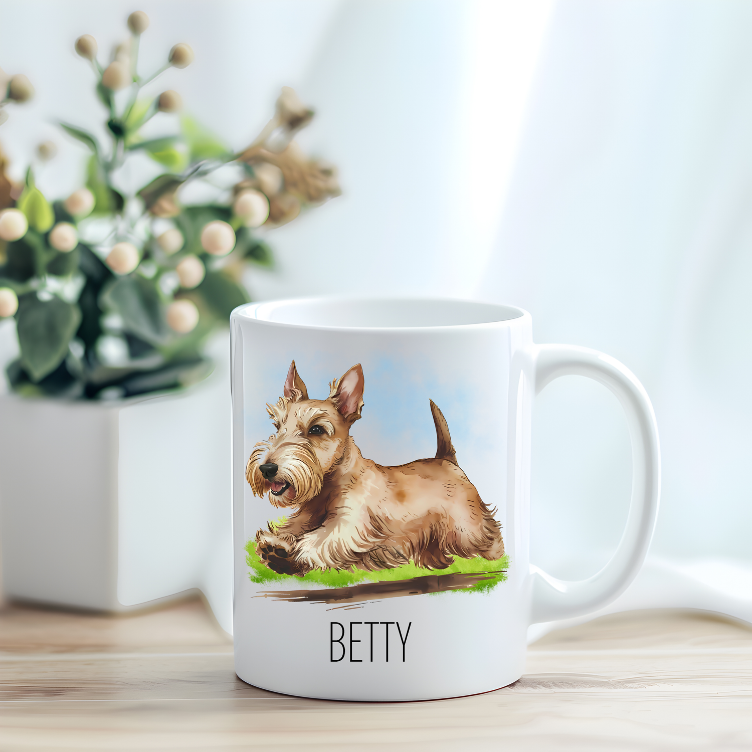 This coffee mug features a wheaten Scottish Terrier on a white ceramic mug which has been placed on a light wooden surface.  A green and white plant sits on the left side of the image in a square plant pot.