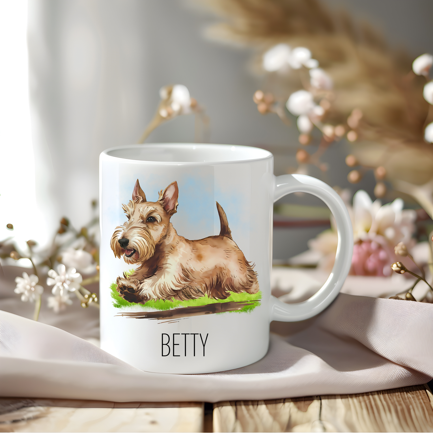 A lovely wheaten Scottish Terrier coffee mug personalised with a pet name.  The mug is sat on a wooden surface with a creamy coloured cloth and a variety of flowers decorate the background.

