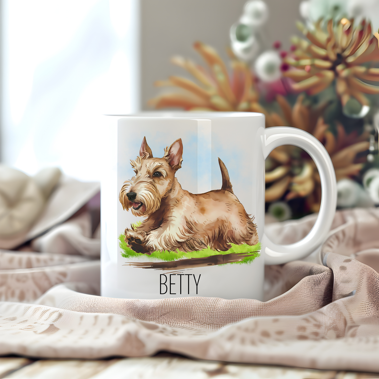 A white ceramic mug with a vibrant image of a cute wheaten Scottish Terrier sits on a beige tablecloth placed on a wooden surface.  The mug is personalised with a pet name and stems of large flowers decorate the background.

