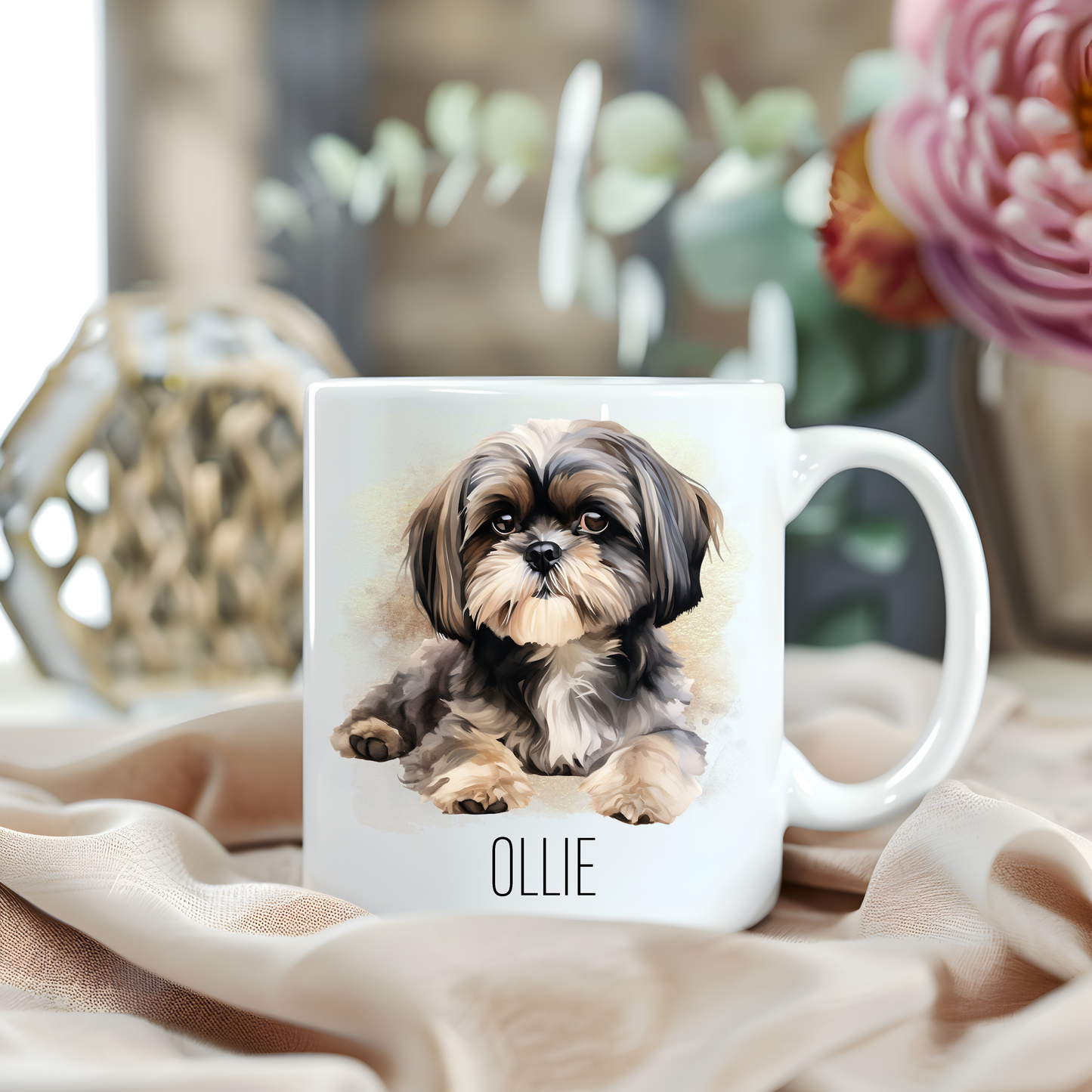 A Shih Tzu laid looking forward is placed on a white personalised ceramic mug.  The mug sits on a beige cloth with a large pink flower and green foliage accessorizing the background.