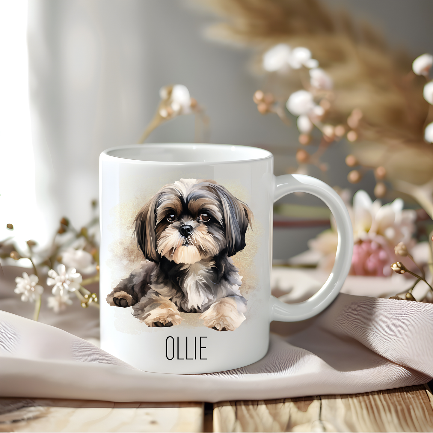 A lovely Shih Tzu coffee mug personalised with a pet name.  The mug is sat on a wooden surface with a creamy coloured cloth and a variety of floral attributes decorate the background.