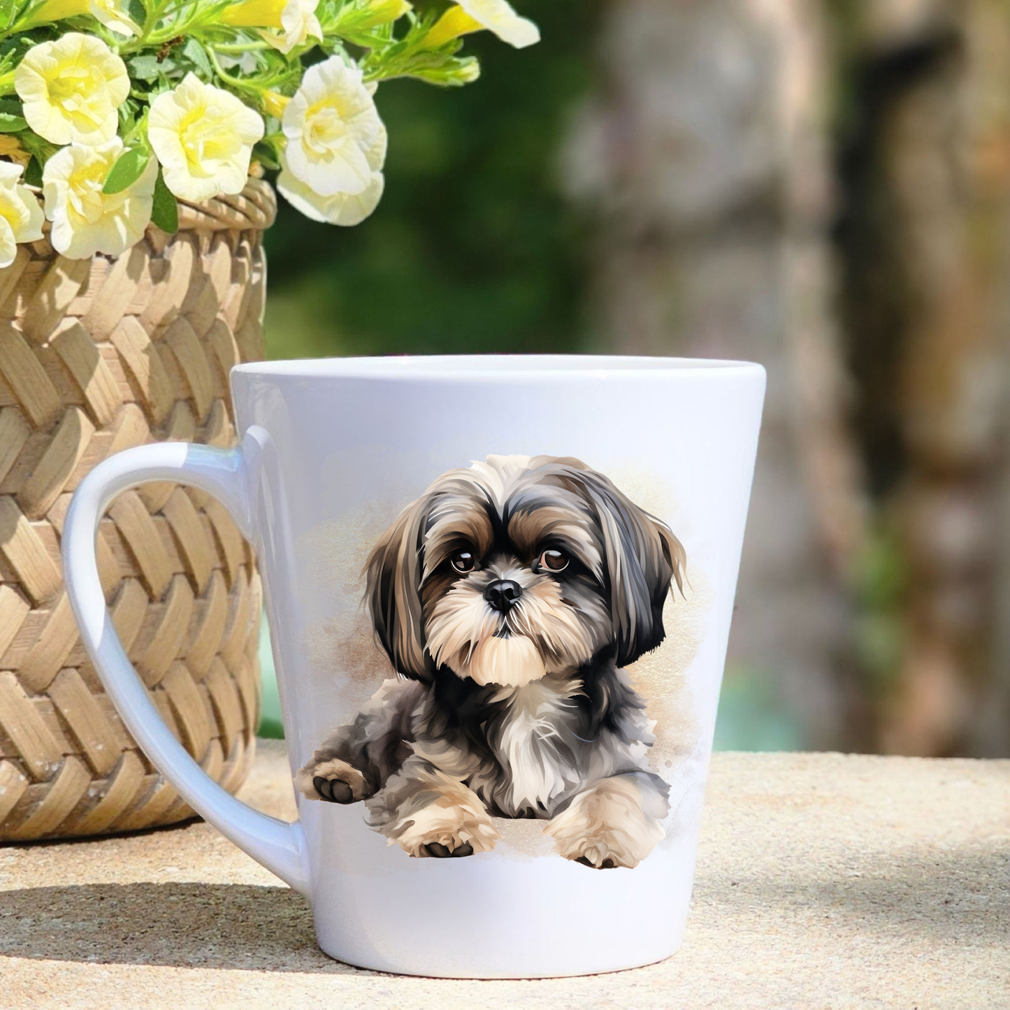 A white ceramic latte mug featuring a Shih Tzu is laid on surface.  A basket containing yellow flowers sits to the left-hand side. 
