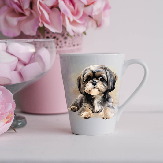 A Shih Tzu is on a white ceramic latte mug. The mug is placed on a white surface with pink flowers in a pink plant pot and a dish of pink marshmallows.