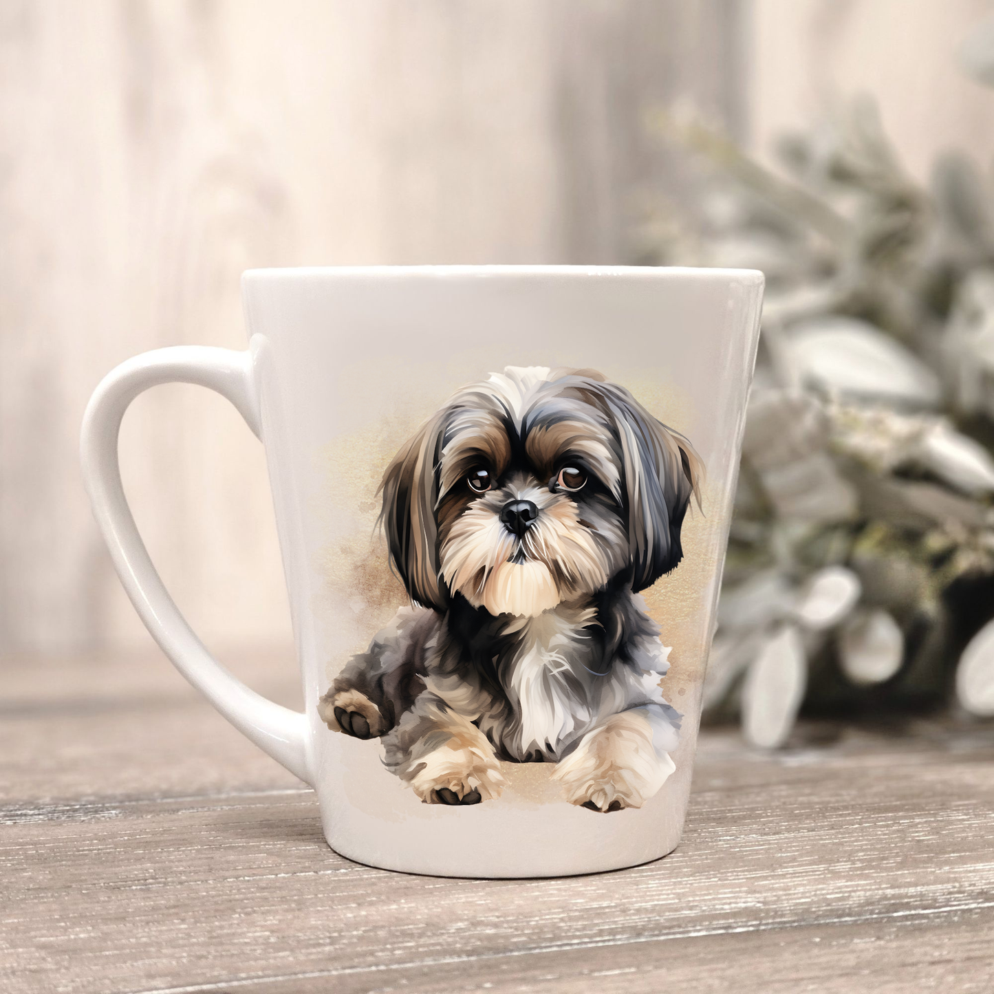 A Shih Tzu is laid on a white ceramic latte mug. The mug is placed on a wooden surface with sage green foliage to the right-hand side.