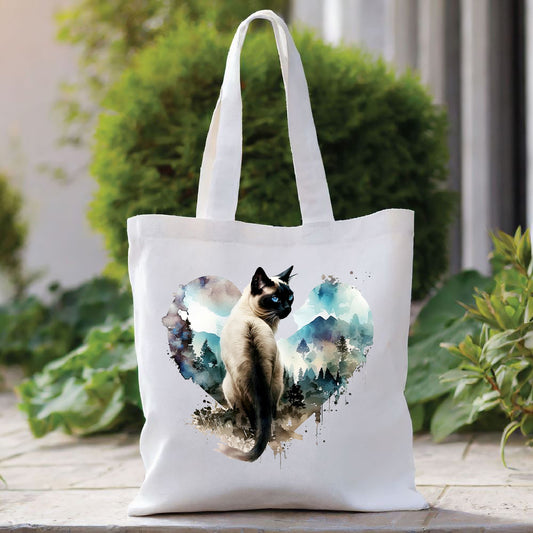 white tote bag showing a siamese cat looking backwards and situated within a blue heart depicting a mountain scene.
