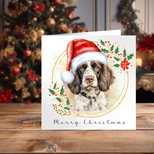 White Christmas Card featuring an English Springer Spaniel wearing a red Santa Hat. Card placed in front of a decorated Christmas tree.
