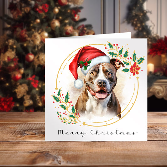 Christmas themed card featuring a Staffordshire Bull Terrier wearing a red Santa hat. Card on a wooden surface and a Christmas tree sits behind.