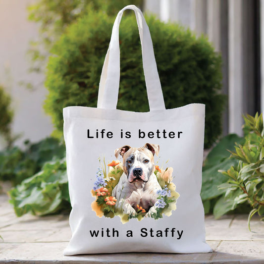 White Tote Bag with the image of a parti-colour Staffordshire Bull Terrier laid among flowers and looking forward. A quote decorates the bag top and bottom. A green bush sits behind with foliage to the right