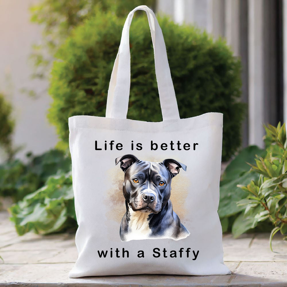 White Tote Bag with a head shot of a black Staffordshire Bull Terrier with a white chest sat gazing forward. A quote decorates the bag top and bottom. A green bush sits behind with foliage to the right