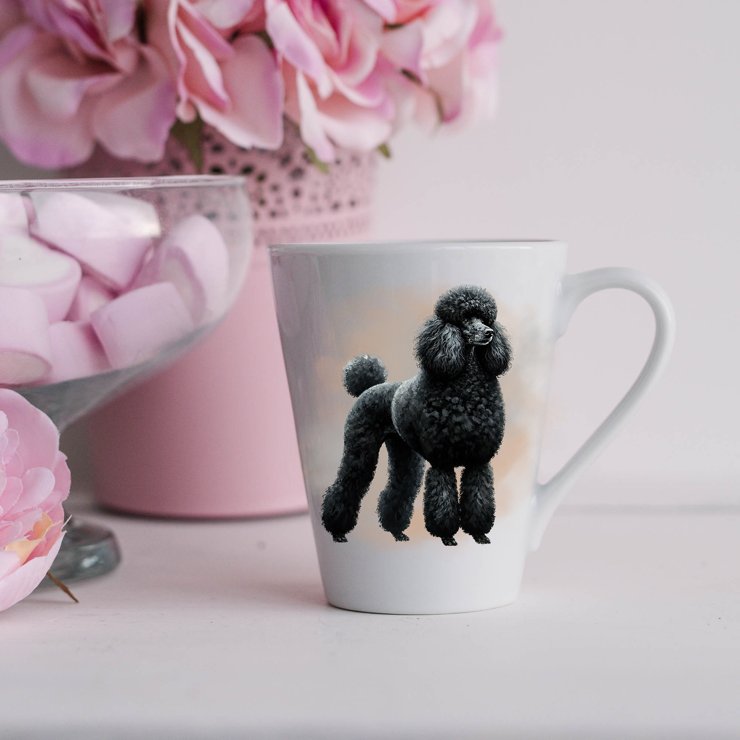 A black Standard Poodle is shown on a white ceramic latte mug in a smoky background and looking forward.  The mug is placed on a white surface with pink flowers in a pink plant pot and a dish of pink marshmallows.
