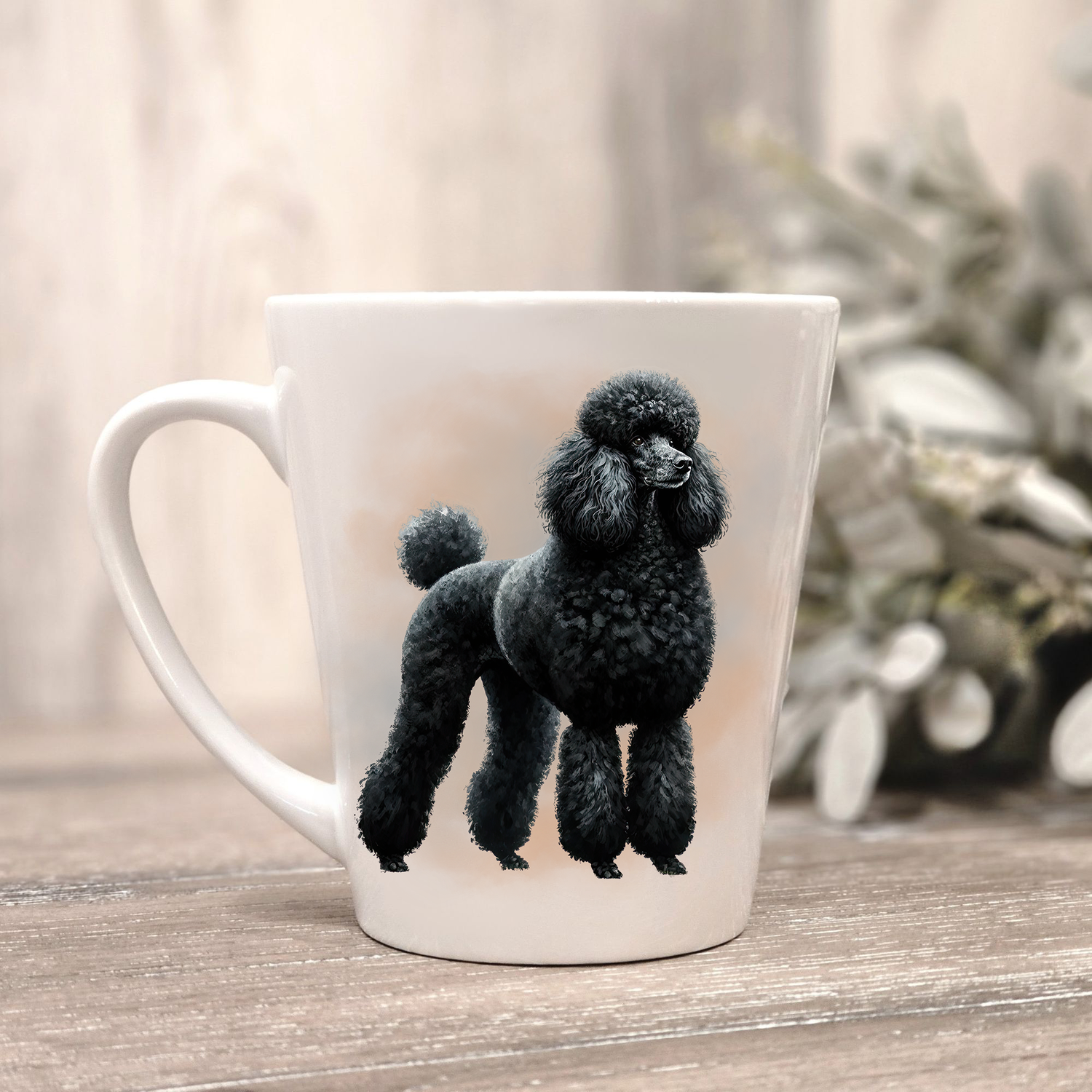 A black Standard Poodle is shown on a white ceramic latte mug in a smoky background and looking to it's left.  The mug is placed on a wooden surface with sage green foliage to the right-hand side.