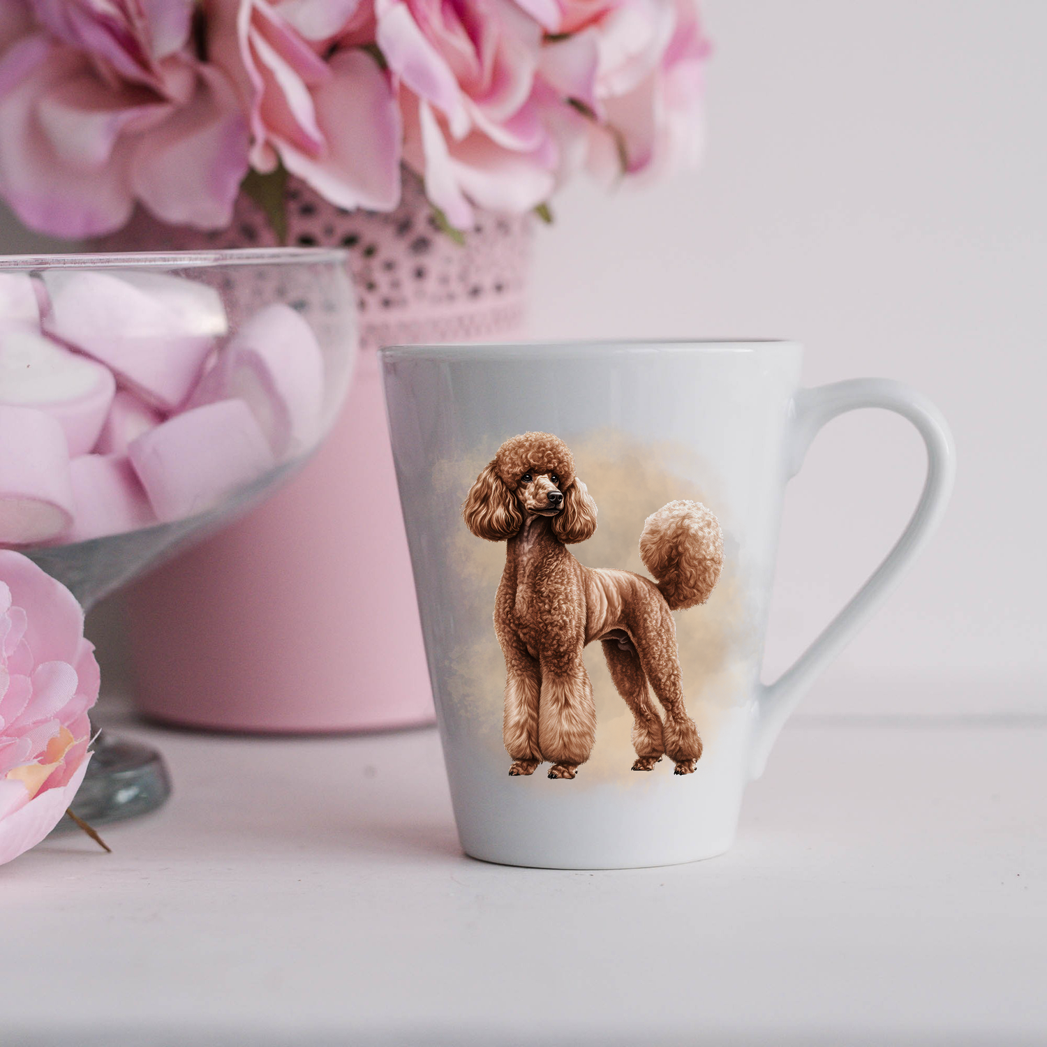 A brown Standard Poodle is shown on a white ceramic latte mug in a smoky background and looking left.  The mug is placed on a white surface with pink flowers in a pink plant pot and a dish of pink marshmallows.