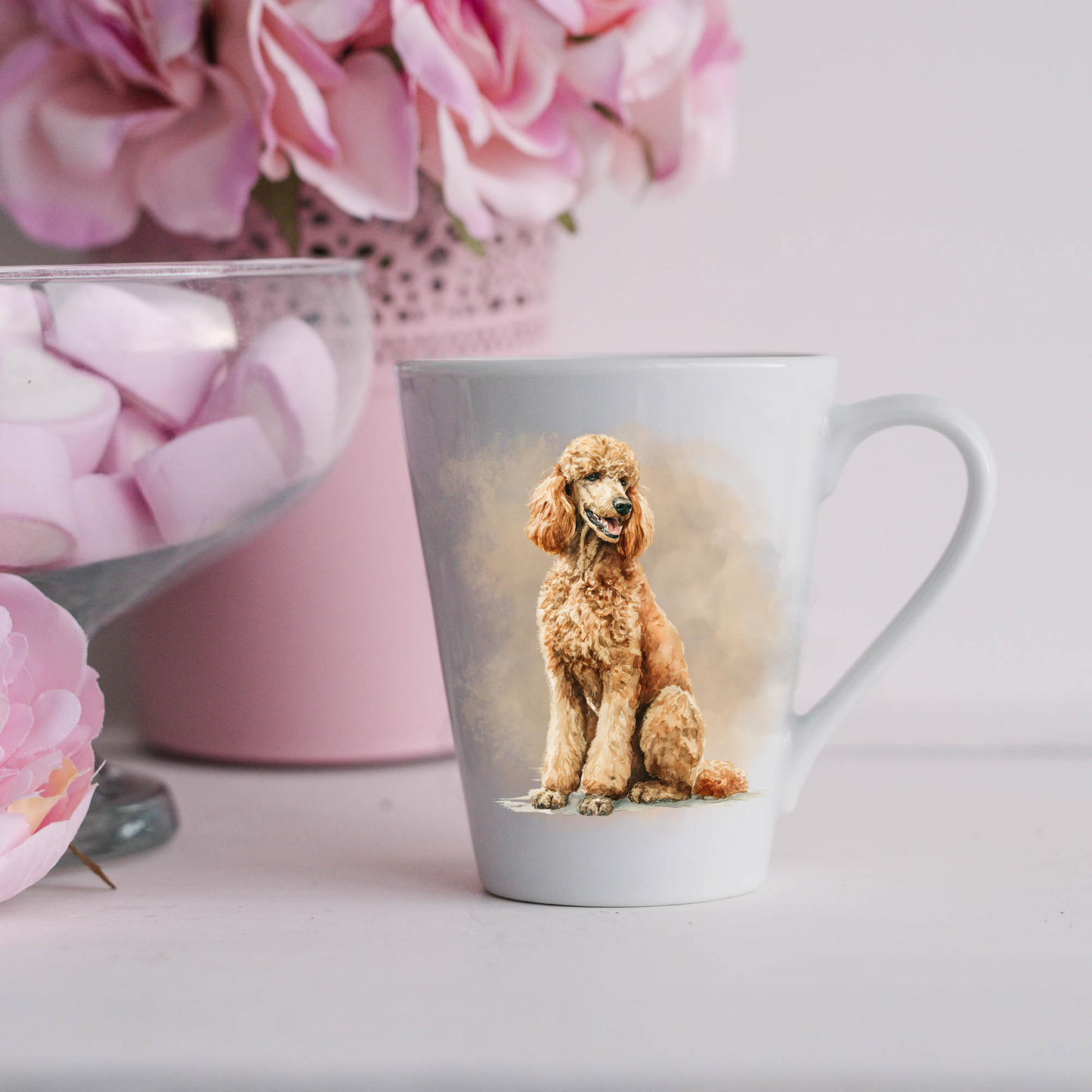 An Apricot Standard Poodle is shown on a white ceramic latte mug in a smoky background and looking to its left.  The mug is placed on a white surface with pink flowers in a pink plant pot and a dish of pink marshmallows.