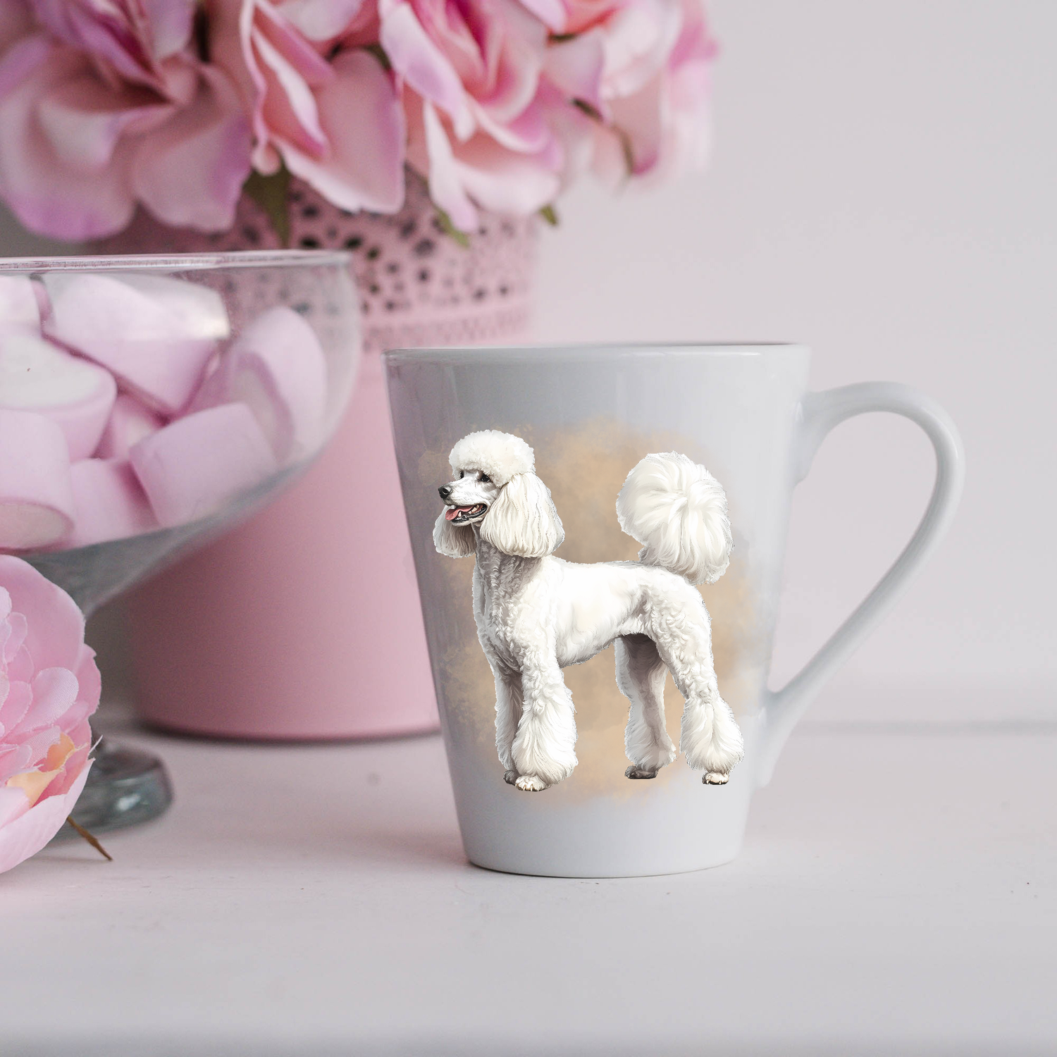 A white Standard Poodle is shown on a white ceramic latte mug in a smoky background and looking to its right.  The mug is placed on a white surface with pink flowers in a pink plant pot and a dish of pink marshmallows.