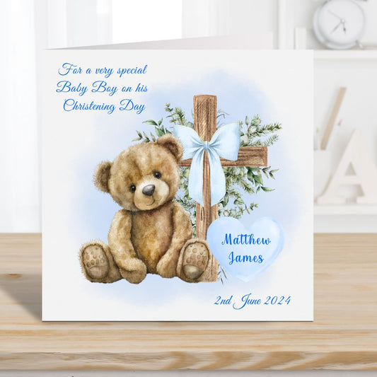 Personalised Christening card for a boy. Teddy bear and cross with a blue ribbon bow. Card placed on a wooden surface with Alphabet letters and a clock in the background.