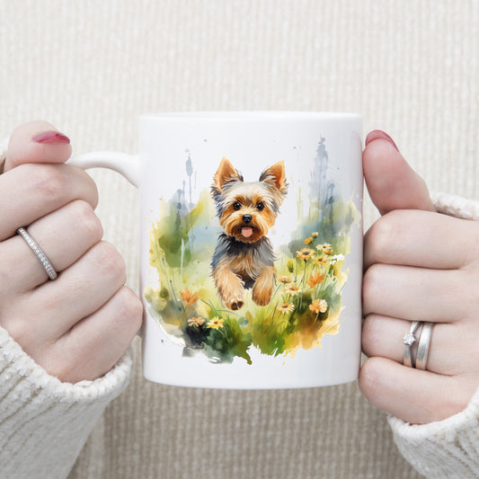 Yorkshire Terrier dog with mouth open and tongue out is running through grass and orange flowers on a white ceramic mug. The mug is being held by a woman with both hands.
