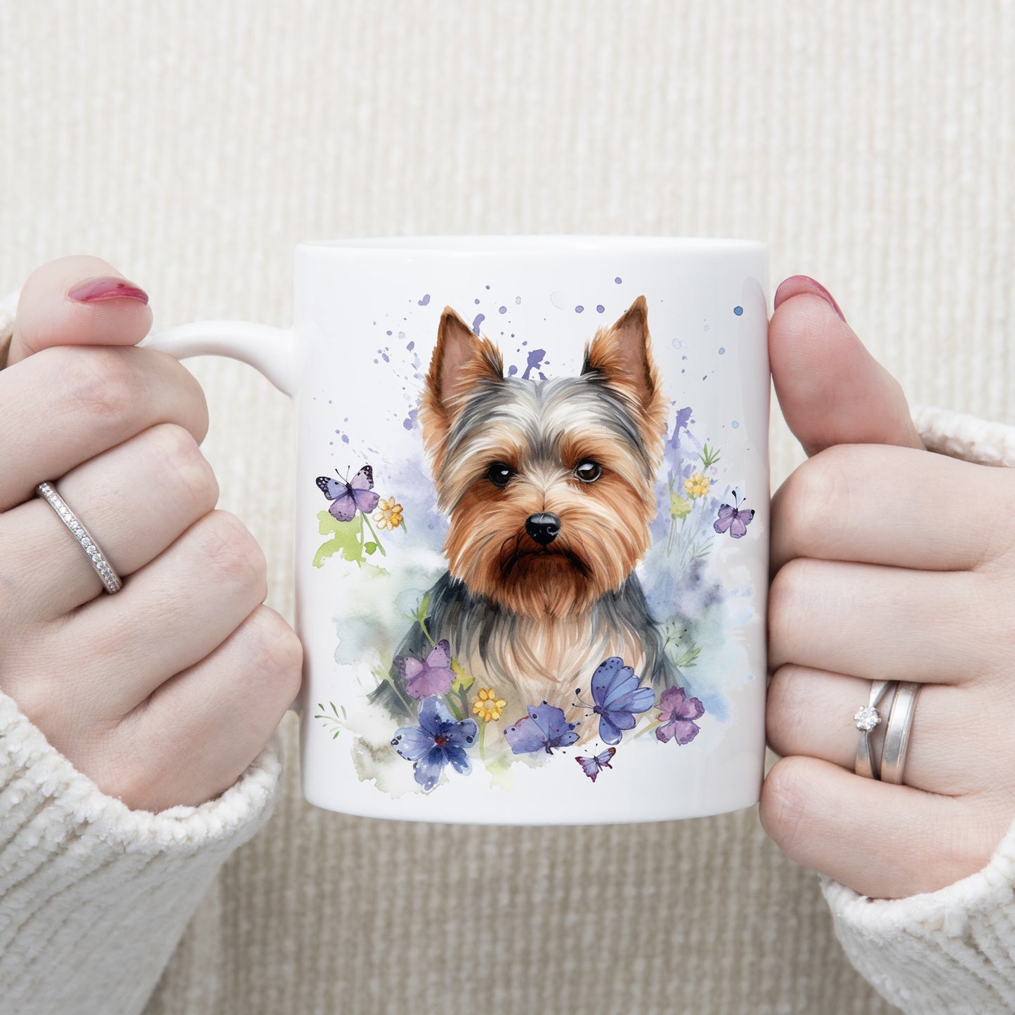 Yorkshire Terrier dog sat on a white ceramic mug.  Blue and purple butterflies decorate with yellow flowers and purple splashes.  The mug is being held by a woman with both hands.
