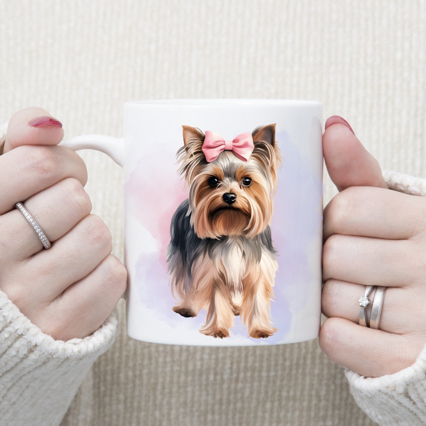 Yorkshire Terrier with a pink ribbon bow on her head is stood facing forward on a white ceramic mug.  A pink and lilac smoky background decorates.  The mug is being held by a woman with both hands.