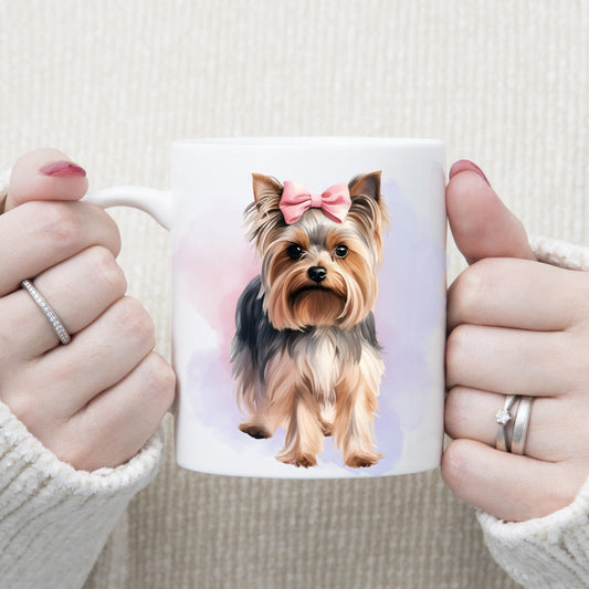 Yorkshire Terrier with a pink ribbon bow on her head is stood facing forward on a white ceramic mug. A pink and lilac smoky background decorates. The mug is being held by a woman with both hands.