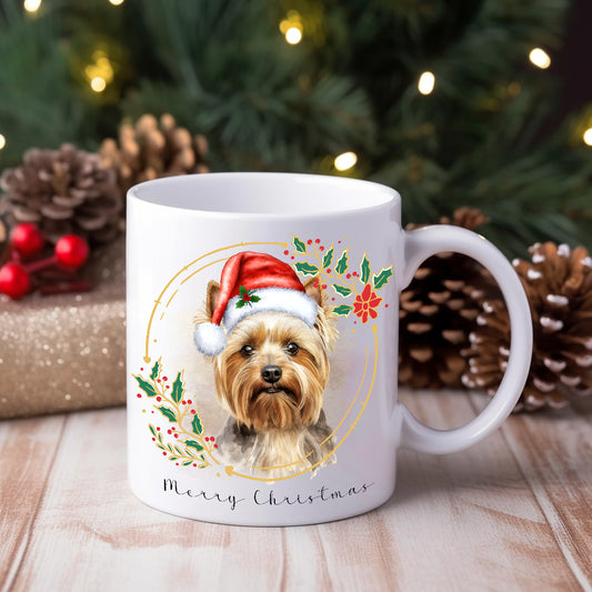 A ceramic mug with a watercolour design featuring a Yorkshire Terrier wearing a Santa hat, positioned in front of a Christmas tree and gifts.
