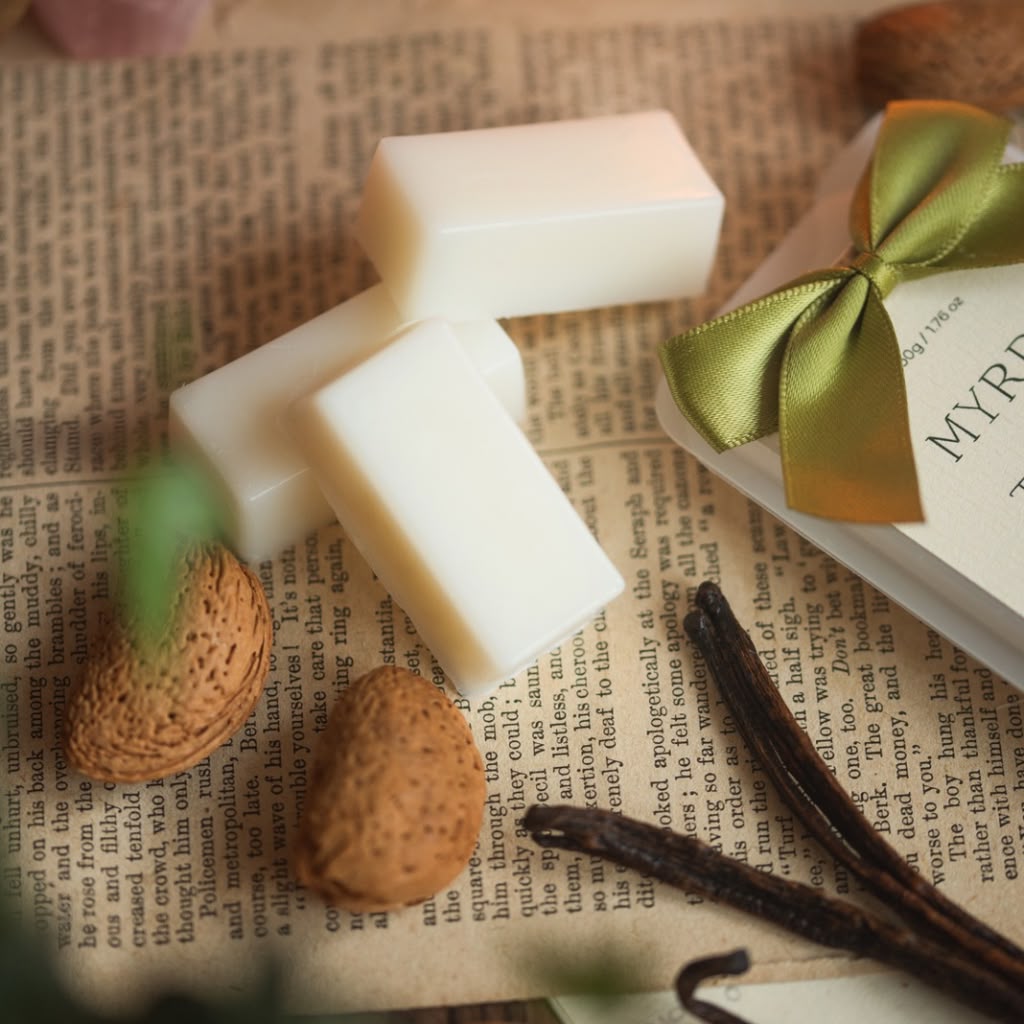 Three white rectangular wax melts or soap bars arranged on top of an open book page, with vanilla beans and whole nuts nearby and a gift box with a gold ribbon at the side.