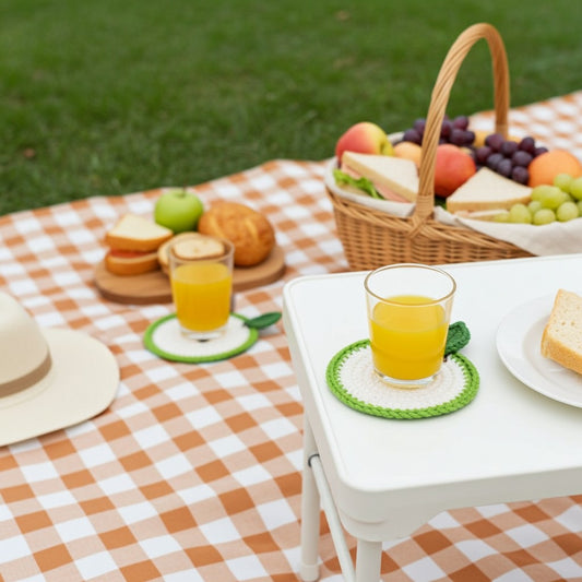 A white folding table set up outdoors on a peach-and-white checkered picnic blanket, with a glass of orange juice resting on a green crocheted coaster. A straw hat, bread, fruit basket, and more food are visible in the background on grass.
