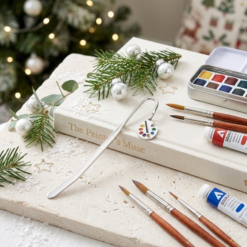 A silver metal bookmark shaped like a paint palette with colourful dots, resting on a cream-colored book titled 'The Painter's Muse'. The scene is styled with Christmas greenery, silver ornaments, paintbrushes, and paint tubes on a stone surface, with a blurred Christmas tree in the background.