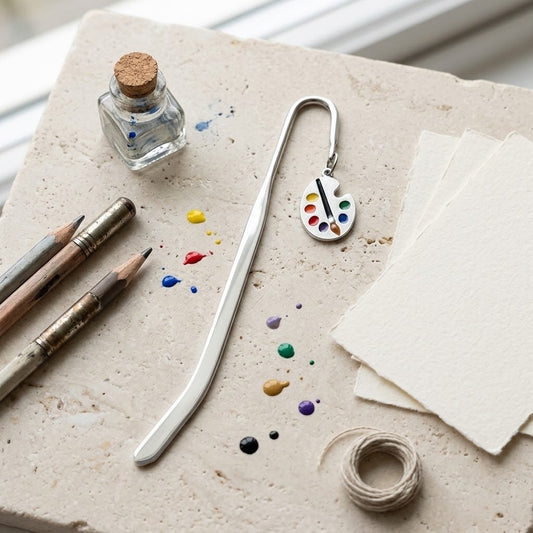 A silver metal bookmark shaped like an artist's paint palette, displayed on a textured stone surface alongside art supplies including paintbrushes, a small glass bottle with a cork, paint splatters, paper, and twine.