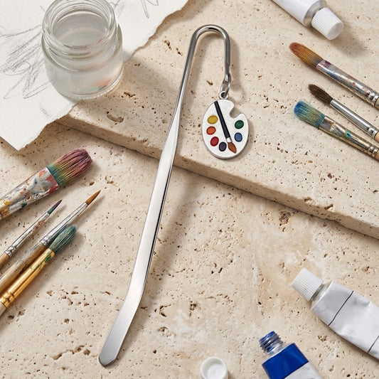 A silver metal bookmark shaped like an artist's paint palette with colourful paint droplets and a paintbrush, displayed on a textured beige stone surface surrounded by art supplies including paintbrushes, tubes, and a small glass jar.