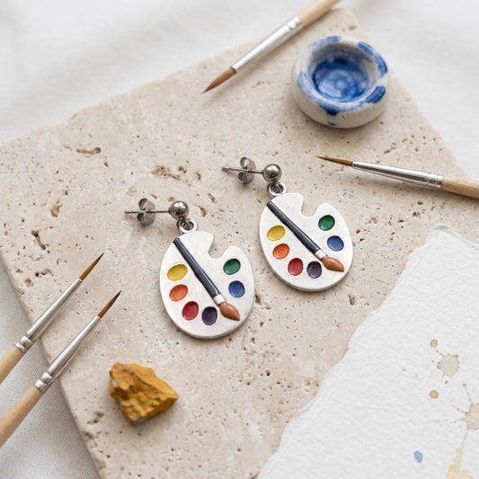 A pair of silver dangle earrings shaped like paint palettes with colourful paint wells and a small paintbrush, displayed on a textured stone slab with art supplies.