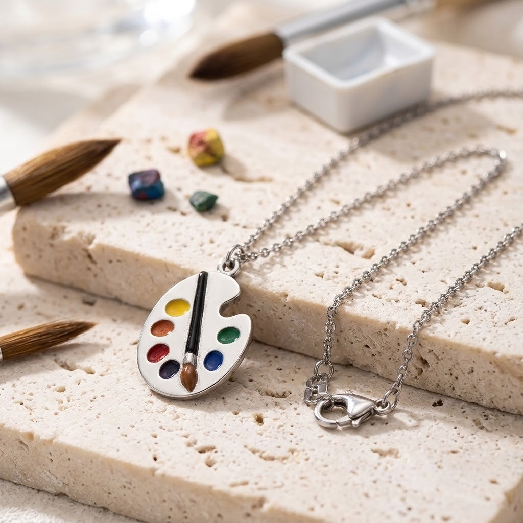 A silver necklace with an artist's paint palette pendant, styled on a textured beige stone slab with paintbrushes, paint cubes, and a small palette dish in the background.
