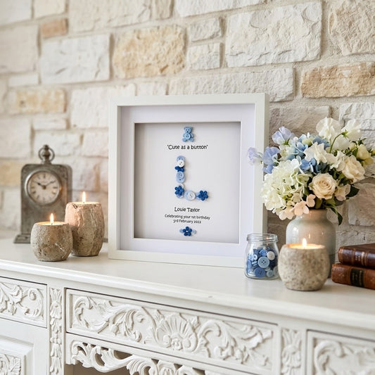 A framed personalised button collage print displayed on a white mantelpiece against a light stone brick wall. The white frame contains blue and white buttons arranged to spell 'Cute as a button' with the text 'Love, Tessa' and a date.
