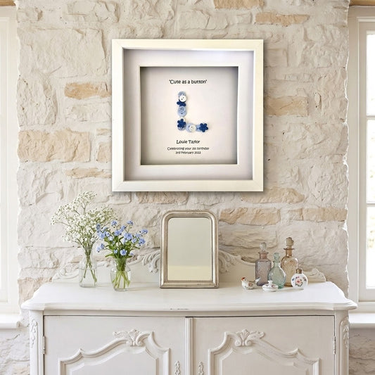 A framed personalised shadow box print hanging on a light stone wall above a white console table. The white frame contains a monogram letter 'L' made from blue and white glass beads with the words 'Cure as a button' and a custom name/date printed below.
