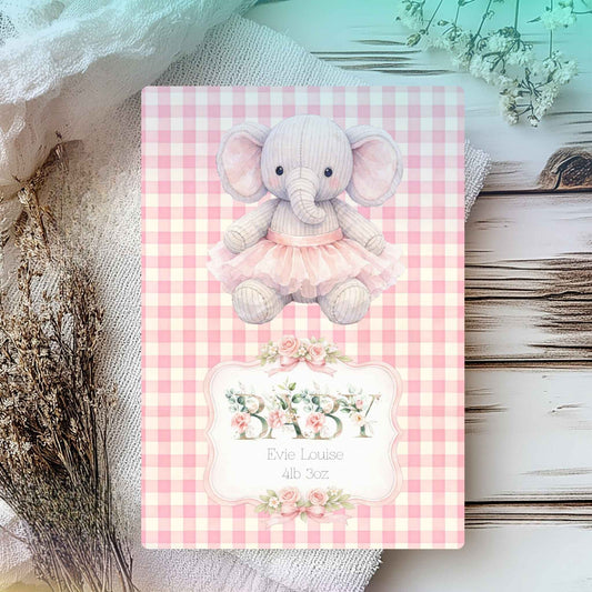A personalised baby card featuring a pink and white gingham pattern, a cute pink tutu elephant, and floral accents. The card is displayed on a rustic white wooden surface surrounded by soft fabric and dried flowers.