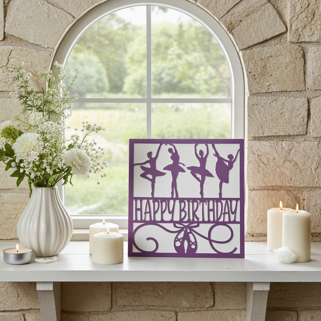 A purple birthday card featuring ballerina silhouettes and the words 'HAPPY BIRTHDAY' stands on a white mantel in front of a stone wall and arched window. White candles, a vase of white flowers, and a small tea light candle are arranged around it.