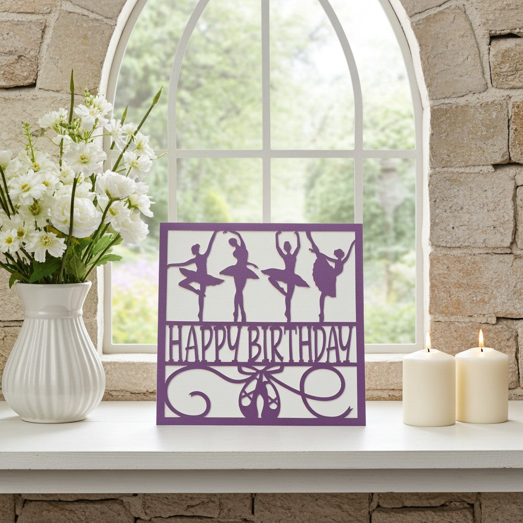 A purple square card featuring ballerina silhouettes and the words 'HAPPY BIRTHDAY' sits on a white windowsill. The sign is styled with a white vase of white flowers on the left and two lit cream pillar candles on the right, set against a stone wall and arched window background.
