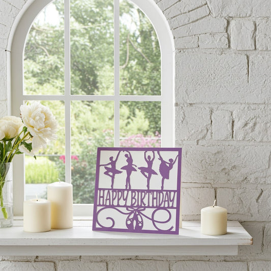 A purple square birthday card featuring ballerina silhouettes and the words 'HAPPY BIRTHDAY' in script, displayed on a white windowsill with cream candles and white flowers against a white brick wall.