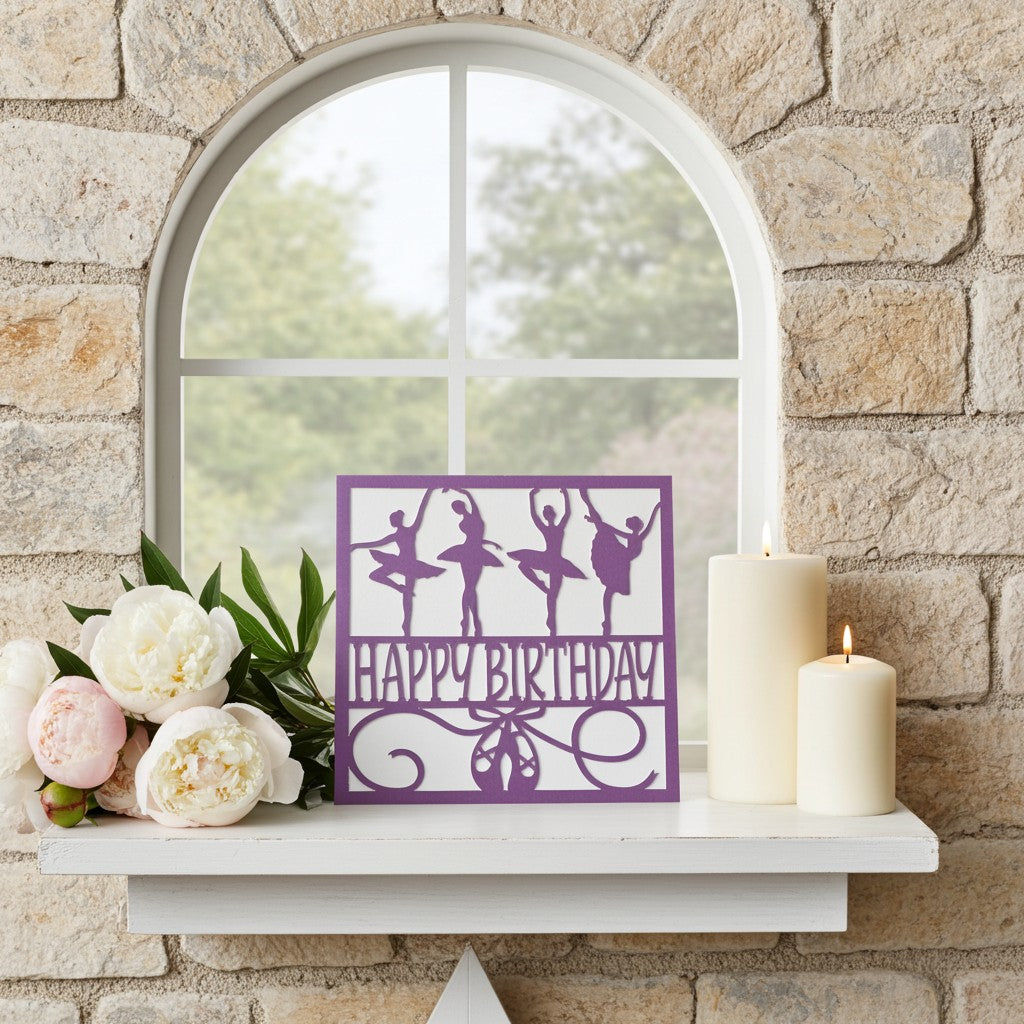 A purple 'Happy Birthday' card featuring ballerina silhouettes, displayed on a white shelf next to white and pink peonies and two lit cream candles against a stone wall and arched window.