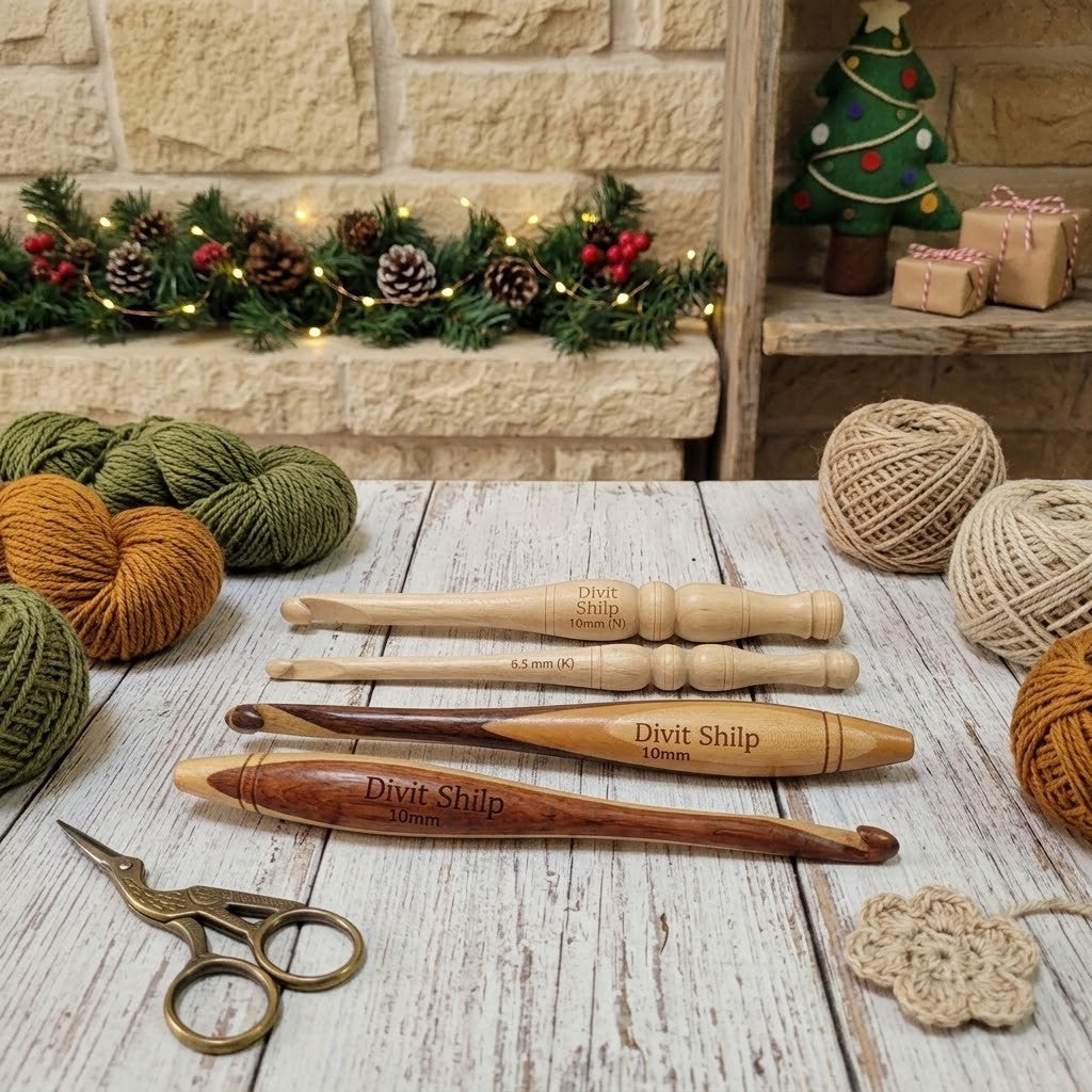 Four Divit Shilp wooden crochet hooks in natural and dark wood finishes arranged on a whitewashed wooden table, with a pair of brass scissors, balls of yarn, and a small crocheted flower nearby. A stone wall and holiday garland are in the background.