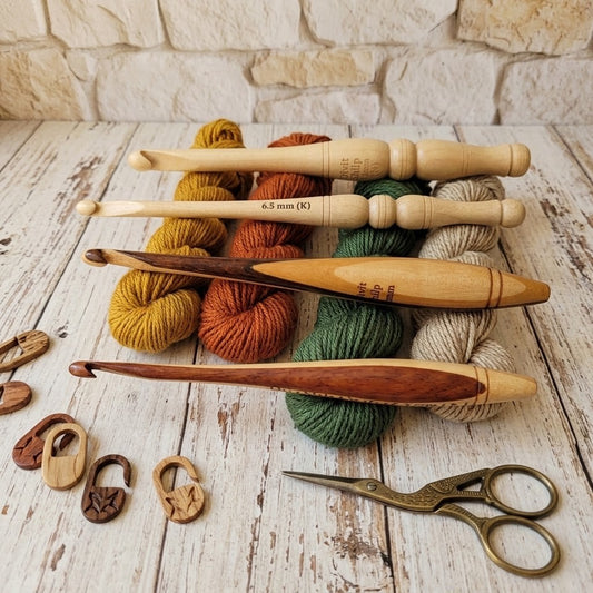 A set of wooden crochet hooks and yarn skeins arranged on a whitewashed wooden surface with a stone wall backdrop. The set includes multiple sizes of wooden crochet hooks, several yarn skeins in mustard yellow, rust orange, sage green, and cream, two wooden stitch markers, and a pair of antique-style metal scissors.