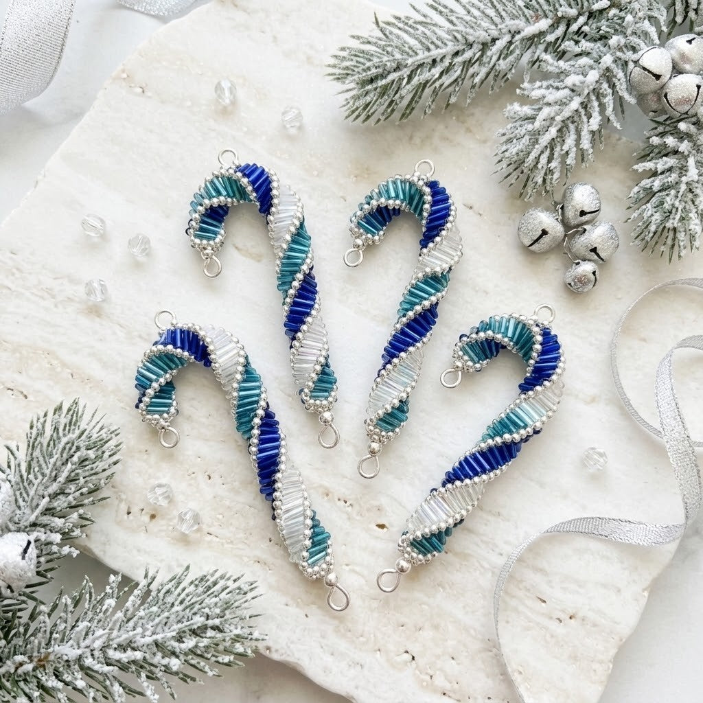 Four beaded candy cane ornaments in blue, teal, and white, arranged on a textured white stone with snowy pine branches, silver bells, and a white ribbon.