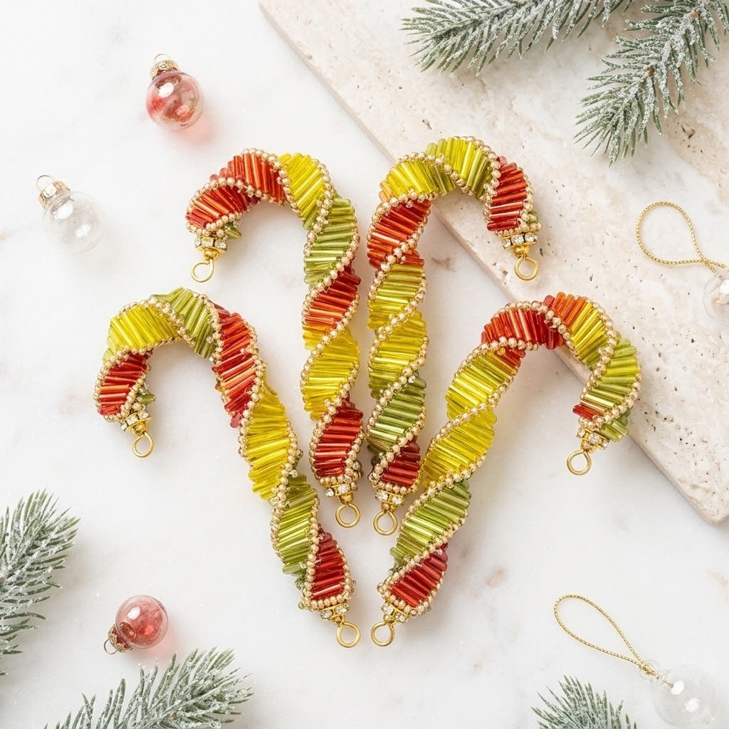 Three handmade beaded candy cane ornaments arranged on a light marble surface with festive greenery and small ornaments around them. The ornaments feature striped red, orange, and yellow sections with white bead accents and gold-tone loops at the tips.