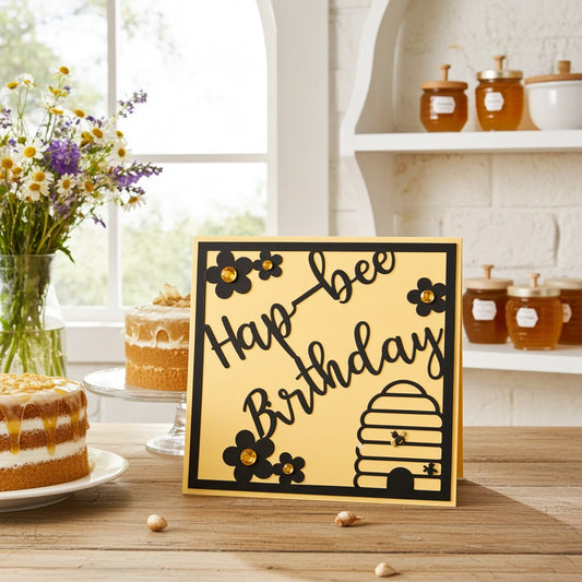 A square greeting card with a yellow background and black border, featuring the words 'Hap-h bee Birthday' in black script with black flower illustrations and a beehive graphic. The card is displayed on a wooden table in a kitchen setting with cakes, honey jars, and flowers in the background.