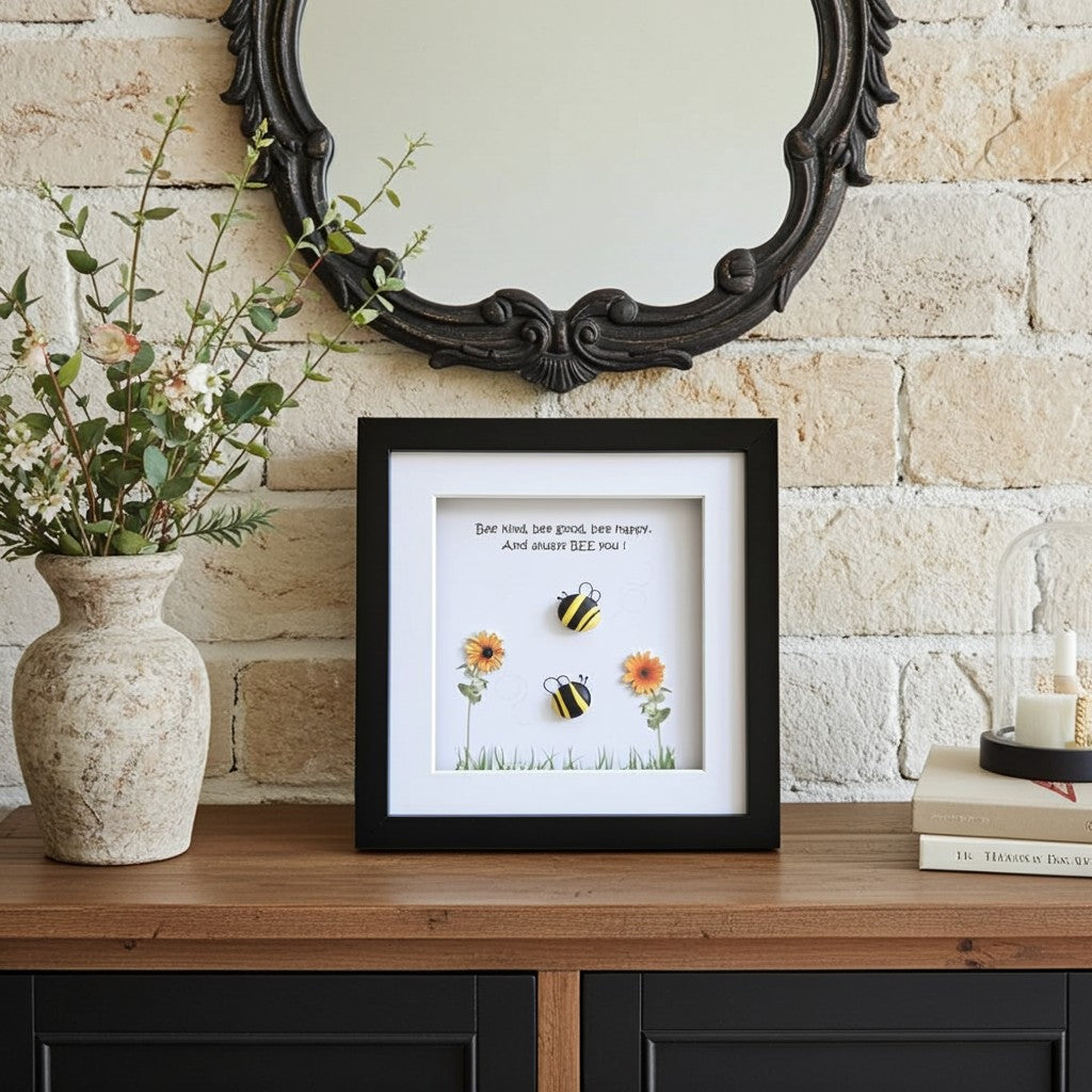 A framed art print featuring two bumblebees and sunflowers with a quote displayed on a wooden console table in a styled room setting with a brick wall, a mirror, a vase, and books.