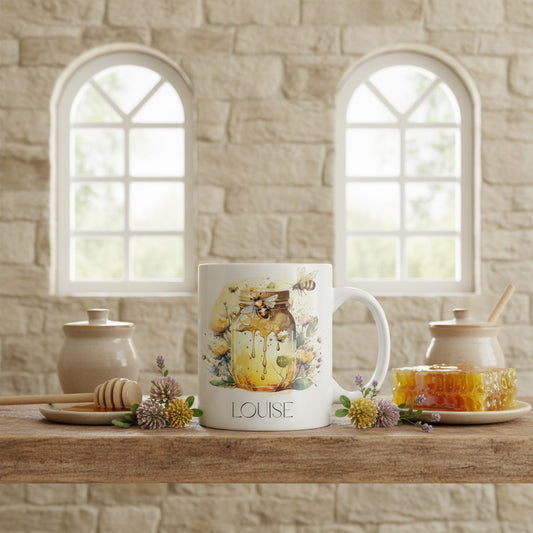 Mug with floral bee design and 'LOUISE' text on a wooden surface with honey jars and flowers.