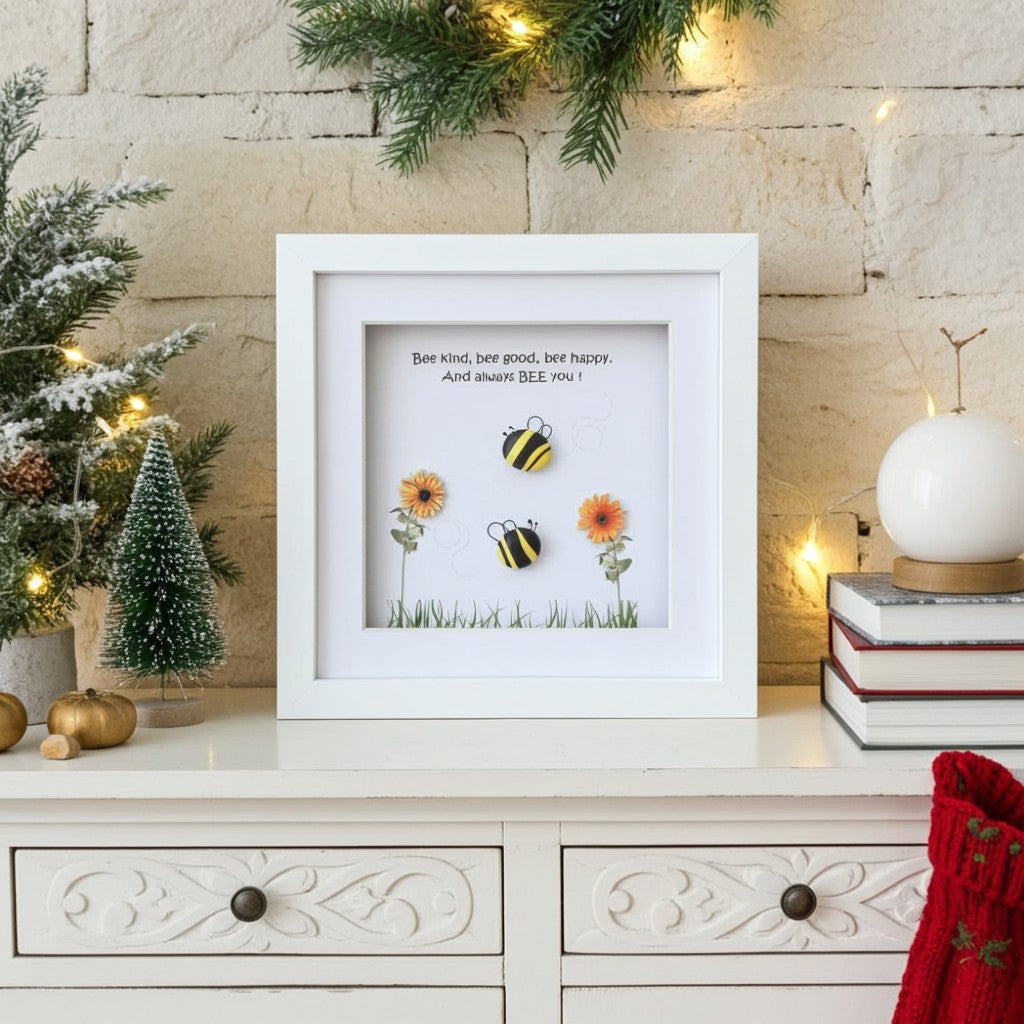 A white framed art print featuring two bumblebees and two orange flowers with the text 'Bee kind, bee good, bee happy. And always BEE you!', displayed on a white cabinet against a rustic stone wall with holiday decorations.